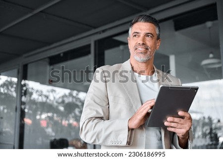 Smiling mid aged business man wearing suit standing outside office holding digital tablet. Mature businessman professional holding fintech device looking away thinking or new business ideas solutions. Royalty-Free Stock Photo #2306186995