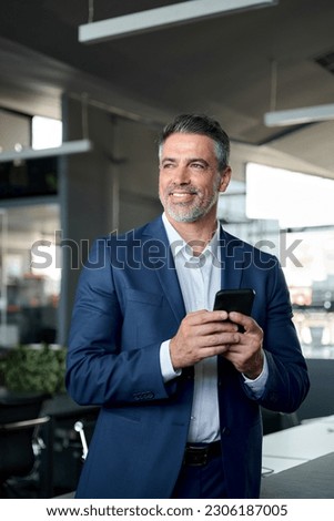 Happy confident mid aged older male company ceo executive wearing suit holding cellphone standing in office using business mobile apps technology financial online solutions on cell phone. Vertical Royalty-Free Stock Photo #2306187005