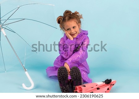 Charming little girl in a purple raincoat and Rubber boots sitting on a ground among umbrellas. A child dressed for a rainy weather. Isolated on blue background.