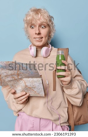 Indoor shot of shocked female tourist focused at paper map with puzzled stunned expression bites lips holds bottle of fresh green smoothie dressed casually poses with rucksack against blue background