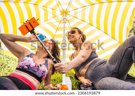 beautiful middle aged women in sportswear having fun taking a selfie under a beach umbrella - healthy lifestyle concept