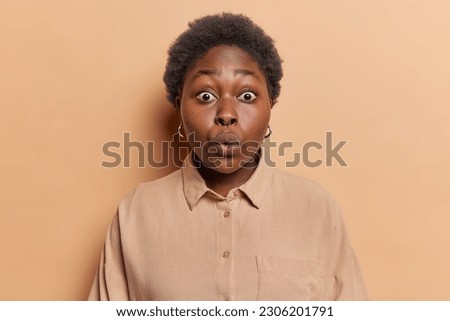 Shocked dark skinned woman stares with omg expression at camera cannot believe own eyes stands speechless reacts to unexpected news wears shirt isolated over brown background. Human face expressions Royalty-Free Stock Photo #2306201791
