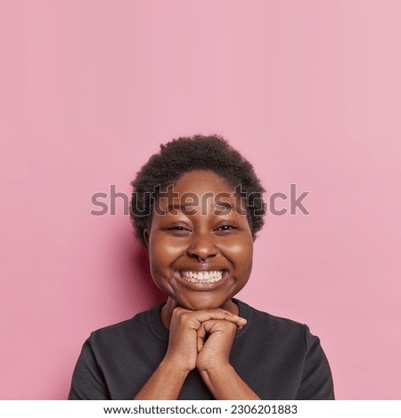 Vertical shot of good looking cheerful African woman keeps hands under chin smiles gladfully looks directly at camera expresses positive emotions dresed casually isolated over pink background Royalty-Free Stock Photo #2306201883