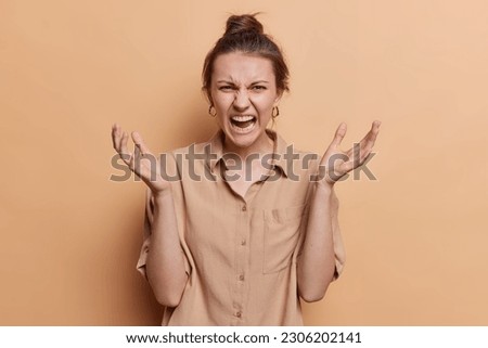 Angry fed up young European woman spreads palms and yells annoyed argues with someone wears shirt expresses negative emotions isolated over brown background. How dare you. Furious female model Royalty-Free Stock Photo #2306202141