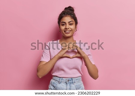 Portrait of happy Indian woman with hair bun shows finger heart gesture sends love to someone smiles pleasantly wears casual clothes isolated on pink background expresses sympathy feels passionate