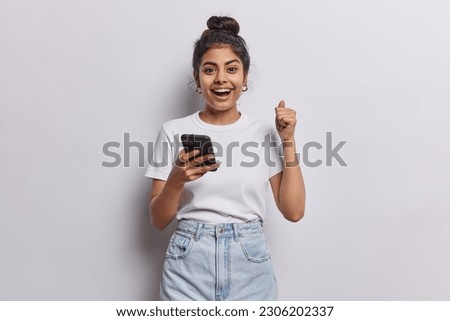 Happy cheerful Indian girl with dark hair combed in bun clenches fist celebrates success holds smartphone dressed in casual basic t shirt jeans isolated over white wall receives message about reward Royalty-Free Stock Photo #2306202337