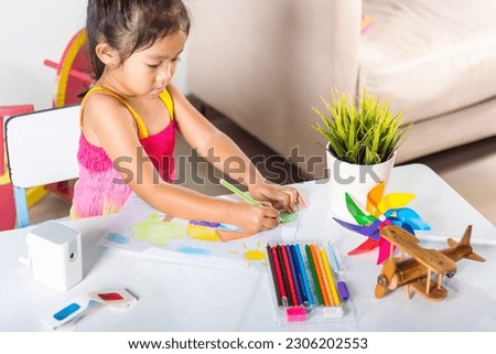 Happy child little girl colorful drawing landscape my home dream on paper, Asian cute kid preschooler sit on table smile she draw country house picture with pencil at home to learning arts homework