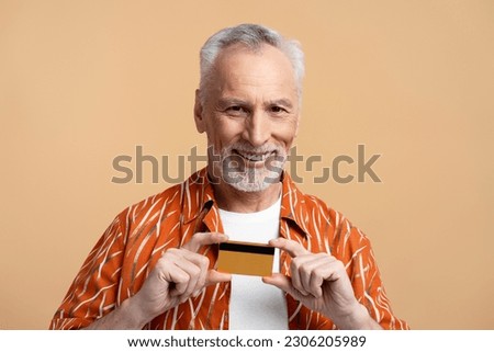 Portrait of smiling senior man holding credit card, looking at camera isolated on beige background. Smiling gray haired pensioner, online banking