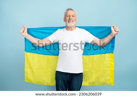 Smiling mature man holding Ukrainian flag isolated on blue background. Attractive male wearing white t shirt mockup, advertisement