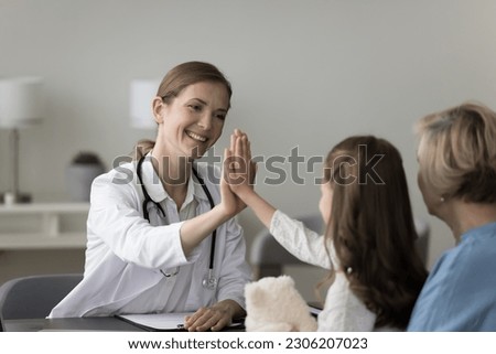 Smiling little girl sit on grandmother laps, give high five to female pediatrician at meeting in clinic, friendly therapist gp greeting welcoming little patient at consultation, celebrate good result Royalty-Free Stock Photo #2306207023