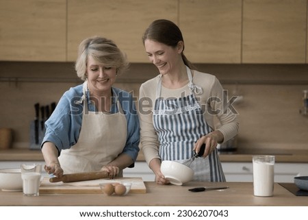 Millennial 35s woman prepare homemade pastries with mature 65s attractive mother wear aprons talking while cooking cake for family holiday in modern kitchen. Family recipe, natural products, cookery Royalty-Free Stock Photo #2306207043