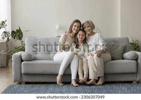 Three generations of women sit on sofa smile look at camera. Elderly 60s grandma her cute little grandchild and grownup daughter shooting for picture in living room. Multigenerational family portrait