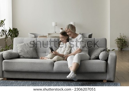 Cheerful grandmother and granddaughter sit on couch, use cellphone together, play on-line games, communicate with relatives through video call. Multi-generational family spend pastime with modern tech