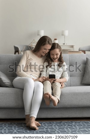 Little girl use mobile phone with young carefree mom sitting on couch at home in living room, looking at screen, smile enjoy new filter, spend leisure time together watch videos with parental control
