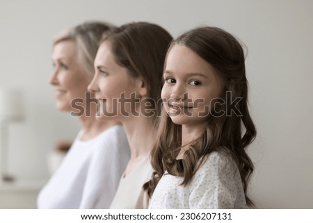 Happy preschooler girl staring at camera posing standing in row with young pretty mother and older grandmother look forward. Multigenerational family shooting indoor. Life from childhood to retirement