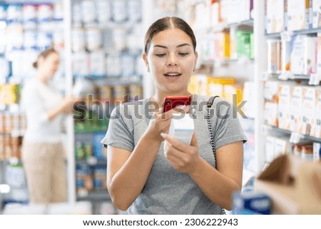 Young woman in casual clothes looking for remedy from photo on internet on smartphone in pharmacy..