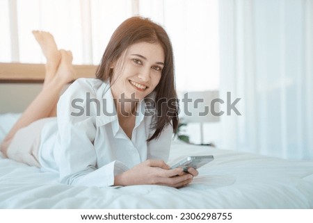 Photo of young happy woman in pajama and holding phone the phone to talk video call Online with friends while sitting on bed.