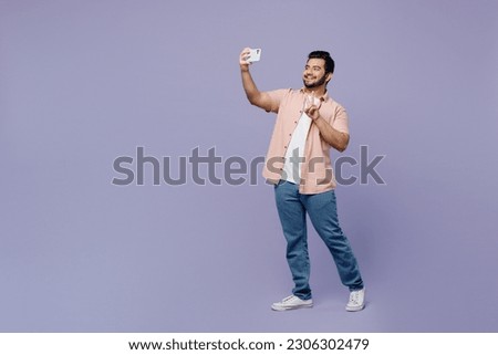 Full body young Indian man he wear pink shirt white t-shirt casual clothes doing selfie shot on mobile cell phone post photo on social network isolated on plain pastel light purple background studio