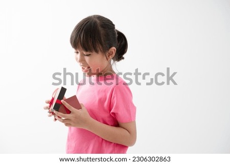 Cute little girl opening her gift, surprised by the contents, looking around and happily smiling, isolated on white background