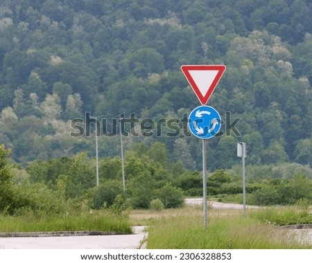 Close-up shot of a yield and roundabout traffic sign attached to a metal pole with forested hills in the background