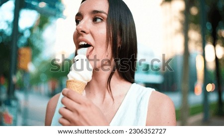 Close-up beautiful woman with freckles and dark loose hair wearing white top testing ice cream. Cute girl enjoys ice cream on modern city background