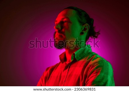 Portrait of man with moustache in casual clothes, hoodie looking at away, posing against dark pink studio background in neon light. Concept of human emotions, facial expression, lifestyle