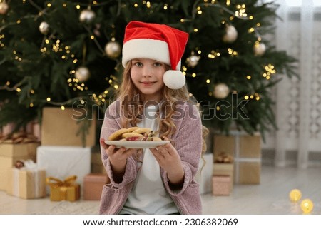 Cute little girl in Santa hat holding plate with cookies near tree and gifts at home. Christmas atmosphere