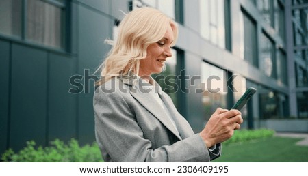 Portrait of happy blond business woman typing by mobile phone outdoors. Woman looking into smartphone screen chatting online or message or social media.