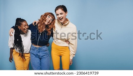 Multiethnic young women laughing and having fun while embracing each other. Group of cheerful female friends enjoying themselves while standing against a blue background. Royalty-Free Stock Photo #2306419739