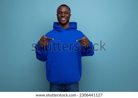 confident dark-skinned young american man in a blue sweatshirt shows his hand at an advertisement on a studio background with copy space