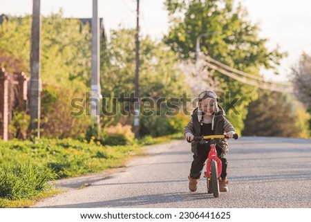 A cheerful little boy rides a bicycle outdoors. A happy child walks in the spring park. The baby is dressed in a fashionable vest, overalls. An aviator's hat is on his head.