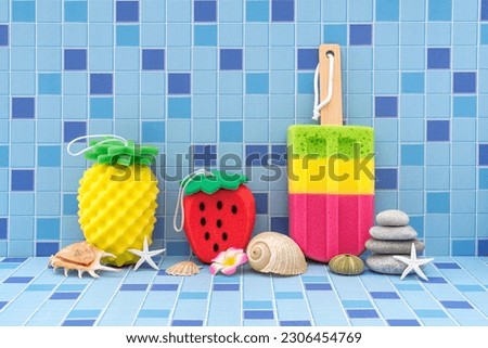 Decorative colored shower sponges in the form of pineapple fruits, strawberries and ice cream on a stick are decorated with seashells. Body care. Blue tile background.