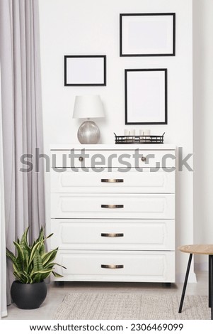 Empty frames hanging on white wall, chest of drawers and potted plant indoors