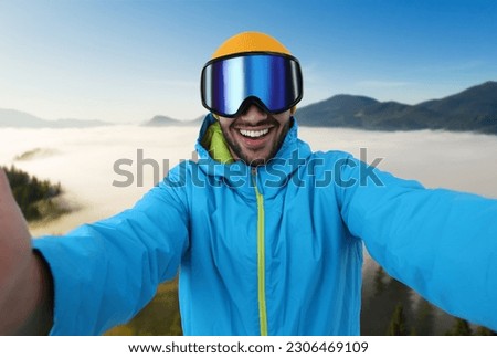 Smiling young man in ski goggles taking selfie in mountains