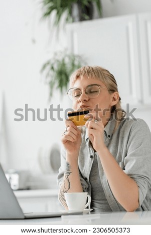young woman in eyeglasses with tattoo on hand kissing credit card, sitting near laptop and cup of coffee on white table, blurred background, work from home, online transactions, technology