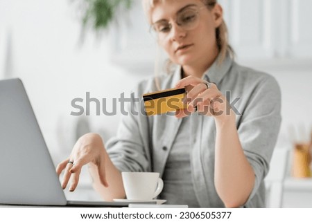 young woman in eyeglasses with tattoo on hand holding credit card, sitting near laptop and cup of coffee on white table, blurred background, work from home, online transactions, technology