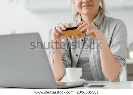 cropped view of happy young woman with tattoo on hand holding credit card, sitting near laptop, smartphone and cup of coffee on white table, blurred background, work from home