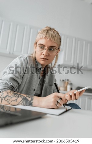 young woman with tattoo on hand writing in notebook, taking notes, holding smartphone and pen near laptop on white table, blurred foreground, work from home