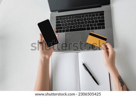 top view of tattooed woman holding credit card and smartphone with blank screen near laptop and notebook with pen on white table, modern workspace, work from home,  digital payment