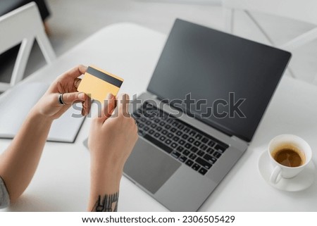 high angle view of tattooed woman holding credit card near blurred laptop with blank screen, notebook and cup of coffee with saucer on white table, modern workspace, work from home, digital payment