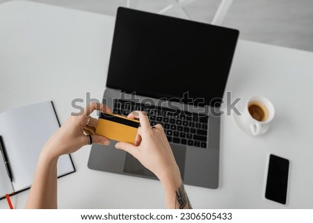 top view of tattooed woman holding credit card near laptop with blank screen, smartphone, notebook with pen, and cup of coffee with saucer on white table, modern workspace, work from home