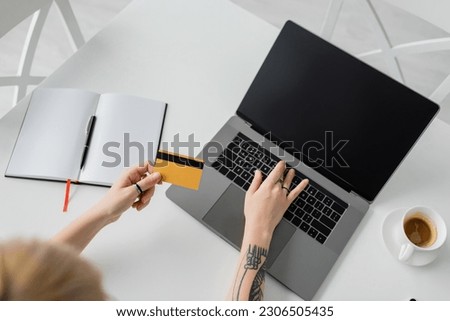 top view of tattooed young woman holding credit card and using laptop with blank screen near notebook with pen, and cup of coffee with saucer on white table, modern workspace, work from home