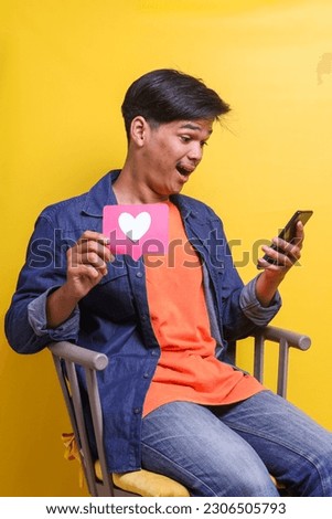 Young surprised Asian man sitting on the chair holding love sign for social media while using mobile phone isolated on yellow background