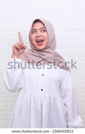 Beautiful Asian Muslim woman in white gamis and hijab pointing finger up and feels joyful isolated over white background. Expression and gesture concept. 