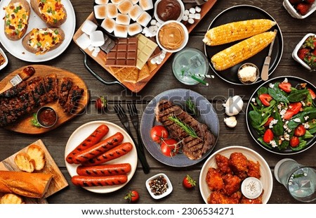 Summer BBQ food table scene over a white wood banner background. Selection of grilled meats, vegetable dishes and smores platter. Overhead view.