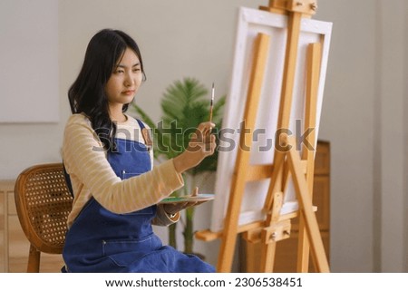 Young woman artist measuring proportion with brush to painting picture in canvas on easel at studio.
