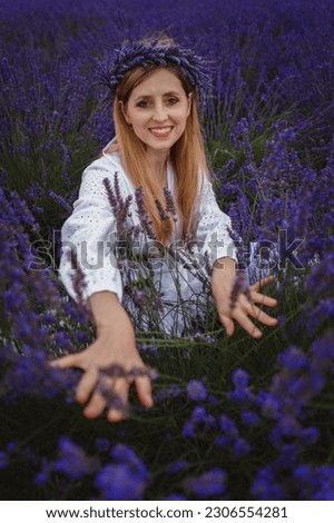 In the picture, a smiling woman, wearing a head wreath, is spreading lavender bushes with the tips of her fingers