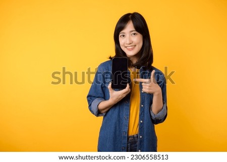 portrait happy young woman wearing yellow t-shirt and denim shirt holding mobile phone and point finger to screen isolated on yellow background. business technology application communication concept.