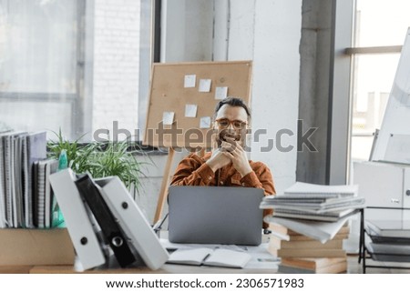 excited businessman in eyeglasses laughing and covering mouth with hands while sitting near laptop, pile of folders, books, notebooks and corkboard with sticky notes on background