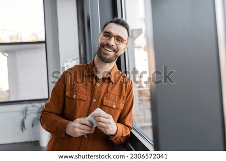 accomplished and charismatic businessman in stylish eyeglasses and shirt standing with mobile phone near window and smiling at camera in office, blurred foreground Royalty-Free Stock Photo #2306572041